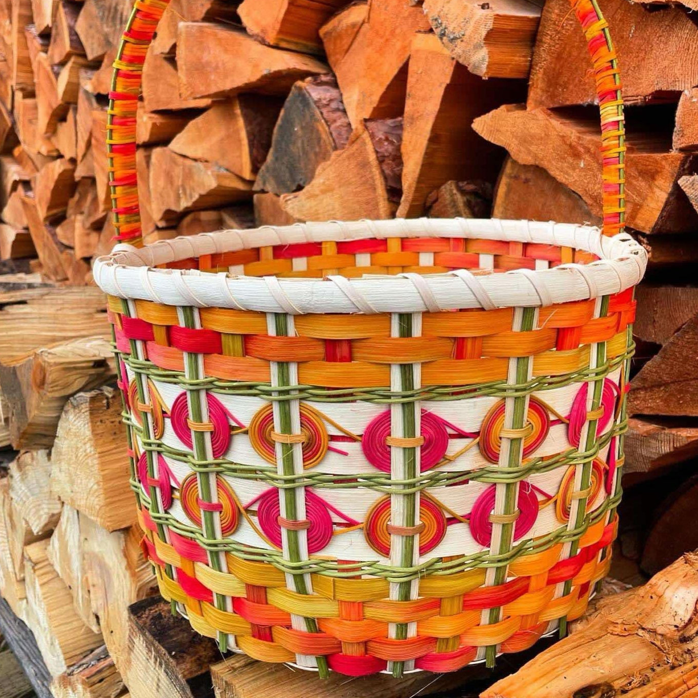 A handcrafted woven basket with intricate patterns in warm hues of orange, red, green, and white, featuring decorative circular embellishments. The basket is set against a neatly stacked pile of firewood, enhancing its rustic charm.